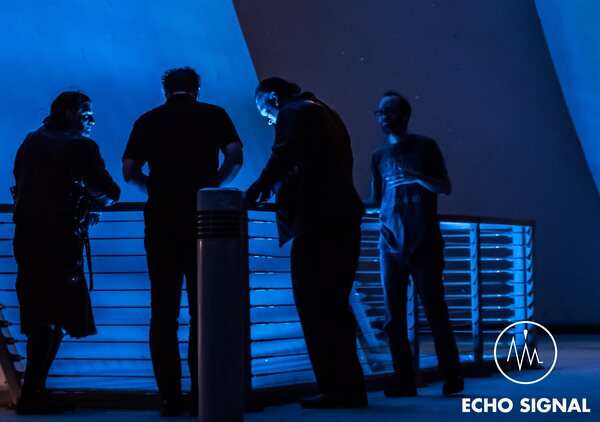 The band photographed from behind, leaning on a railing in heavy blue light with stacked containers beyond.