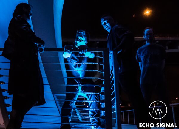 Four members of Echo Signal on an open steel staircase at night, lit hard from below in deep blue, one figure silhouetted in a long coat at the edge of frame.