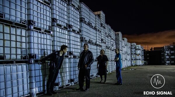 The band spaced along a wall of stacked white industrial containers at night, a band of orange sky at the horizon.