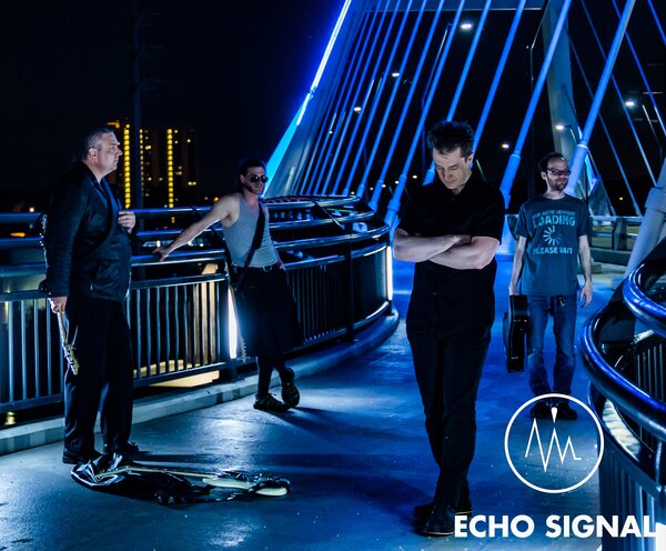 The band spread along a bridge walkway at night, leaning on blue-lit railings with the lights of the Minneapolis skyline behind.