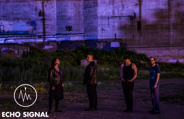 The band standing apart in dry grass in front of a vast concrete grain silo at dusk, the whole scene cast in violet.