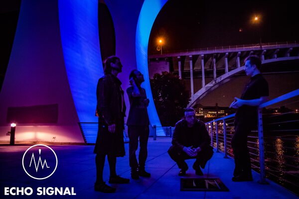 The band standing apart from one another under a bridge span at night, lit in magenta and blue, the lit arch of a second bridge behind them.