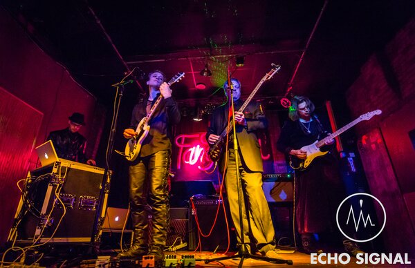 Echo Signal playing live beneath a low concrete ceiling, the stage washed in red with a neon sign behind the amps.
